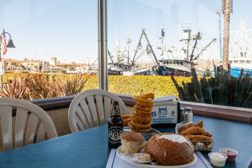 A seaside restaurant table with a meal of fried fish, onion rings, and a drink, overlooking a harbor with fishing boats.