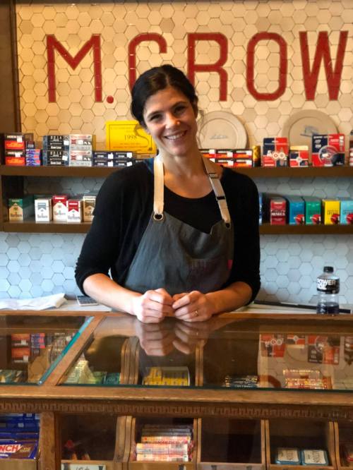 A smiling woman in an apron stands behind a wooden counter filled with various products, with a vintage shop backdrop.