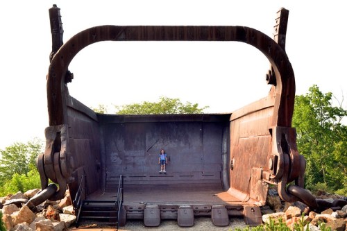 A person stands inside a massive, rusted excavator bucket surrounded by trees and rocks.