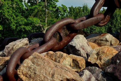 A close-up of a large, rusted chain resting on a pile of colorful rocks, surrounded by greenery.