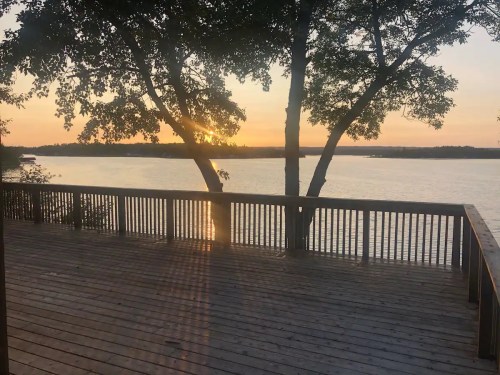 Sunset over a calm lake, viewed from a wooden deck surrounded by trees.