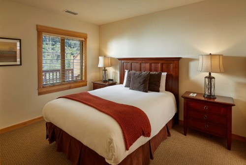 Cozy bedroom with a wooden bed, warm lighting, and a window overlooking greenery. Soft bedding and a bedside table are visible.