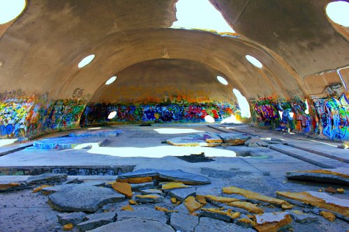 Interior of a graffiti-covered, abandoned building with a domed ceiling and broken concrete floor. Bright light enters through openings.