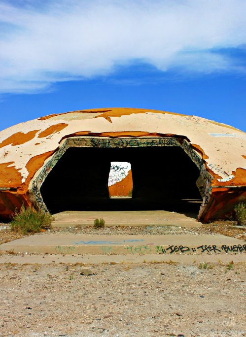 A weathered, dome-shaped structure with graffiti, set against a blue sky and surrounded by sparse vegetation.