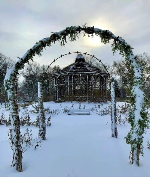 A snowy garden scene featuring a wooden archway leading to a gazebo, surrounded by snow-covered plants.