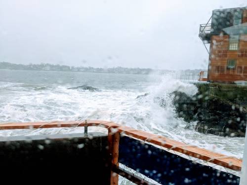 Waves crash against rocks near a wooden structure on a rainy day, viewed through a wet window.
