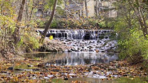 A serene waterfall cascades over rocks, surrounded by autumn foliage and a calm stream reflecting the trees.