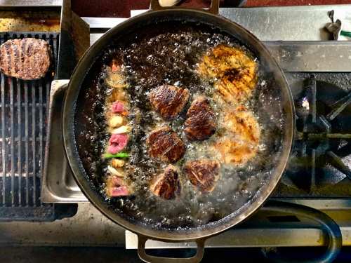A large pot of sizzling meat and vegetables frying in oil on a stove, with a grill in the background.