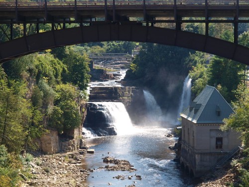 A scenic view of waterfalls beneath a bridge, with lush greenery and a historic building by the water's edge.