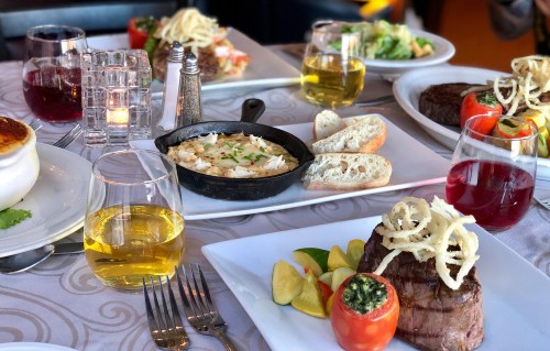 A table set with various gourmet dishes, including steak, seafood, salads, and drinks in elegant glassware.