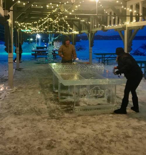 Two people play a game at an ice table under festive lights in a snowy outdoor setting.