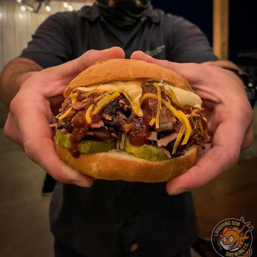 A person holds a loaded sandwich with pulled pork, cheese, pickles, and barbecue sauce, set against a rustic background.