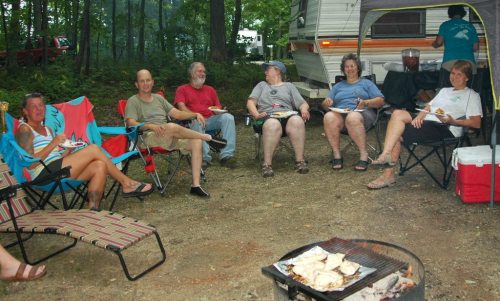 A group of six people sitting around a campfire, enjoying food and conversation near a camper in a wooded area.