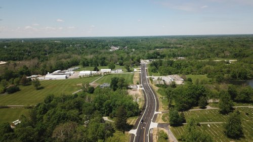 Aerial view of a winding road surrounded by lush greenery and fields, with buildings visible in the distance.