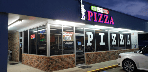 Exterior of New York Pizza restaurant at dusk, featuring large windows and colorful neon signs.