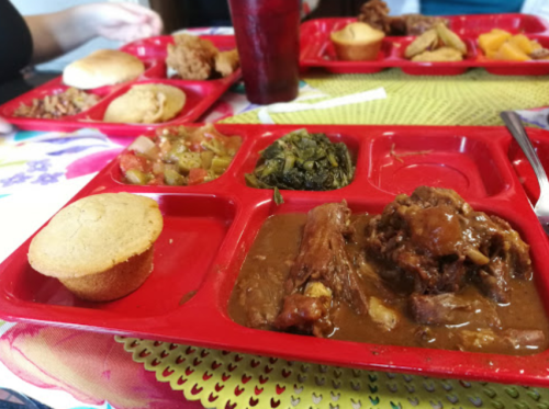 A red cafeteria tray with cornbread, meat in gravy, greens, and a vegetable medley, set on a colorful tablecloth.