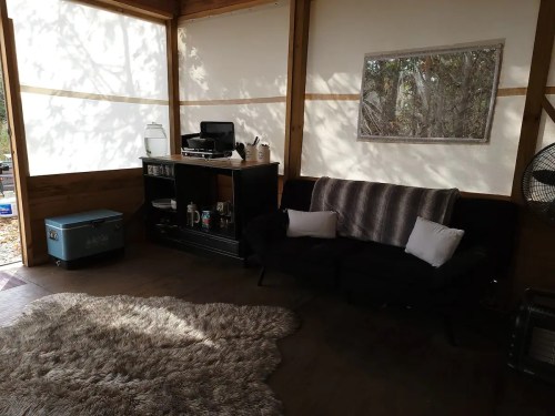 Cozy interior of a cabin with a black couch, a rug, and a wooden cabinet, surrounded by natural light.