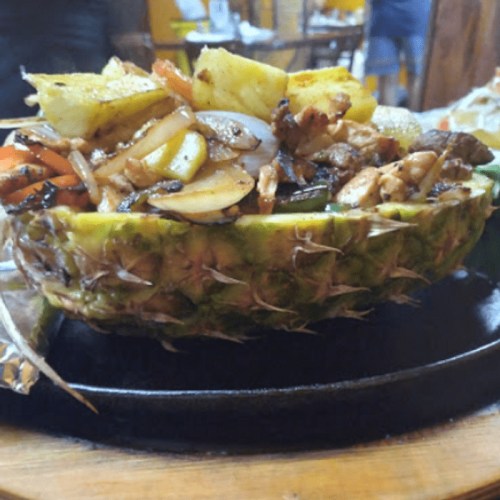 A pineapple half filled with grilled vegetables and meat, served on a sizzling platter.