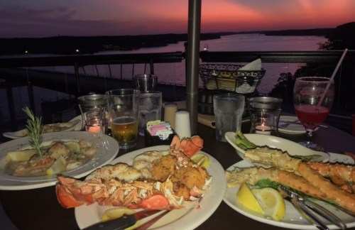 A scenic dinner table with seafood dishes and drinks, overlooking a sunset by the water.