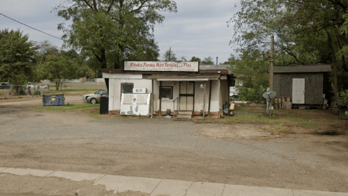 A small, weathered building with a sign reading "Rhonda's Famous Hot Tamales" in a rural area, surrounded by trees and gravel.