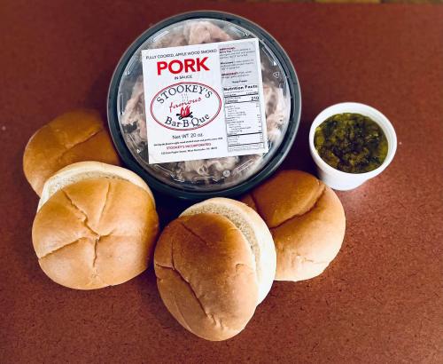 A container of pulled pork with five sandwich buns and a cup of pickles on a wooden surface.