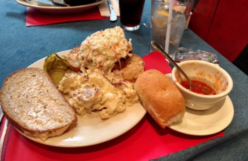 A plate with a sandwich topped with coleslaw, potato salad, pickles, and a small bowl of sauce on the side.