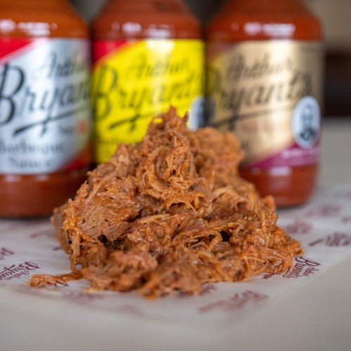 Pulled pork in front of three bottles of Arthur Bryant's barbecue sauce on a paper surface.