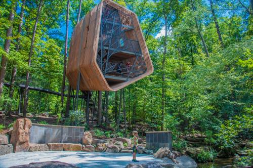 A modern treehouse structure suspended among trees, surrounded by lush greenery and rocky landscaping.