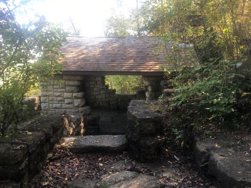 A stone structure with a wooden roof surrounded by trees and foliage in a natural setting.