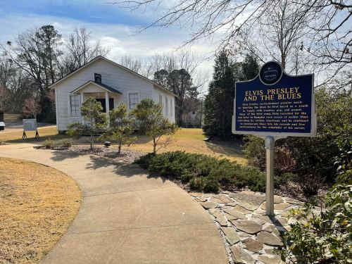 A historic marker about Elvis Presley and the blues, with a white building and landscaped path in the background.