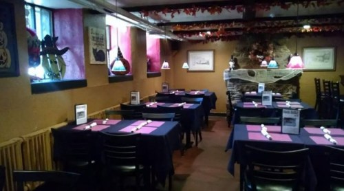 Cozy restaurant interior with dark tables, purple tablecloths, and colorful pendant lights, featuring a stone fireplace.