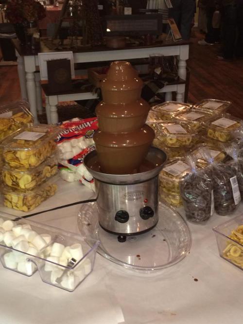 A chocolate fountain surrounded by bags of snacks like marshmallows, pretzels, and cookies on a table.
