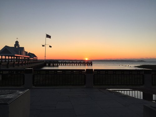 Sunset over a calm waterfront, with a pier and flags silhouetted against the colorful sky.