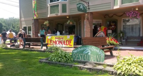 A sunny outdoor scene featuring a restaurant entrance with a lemonade stand and people walking by. Greenery surrounds the area.
