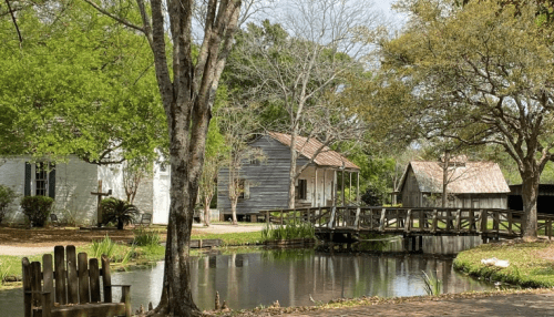A peaceful scene featuring a pond, wooden bridge, and quaint houses surrounded by trees and greenery.