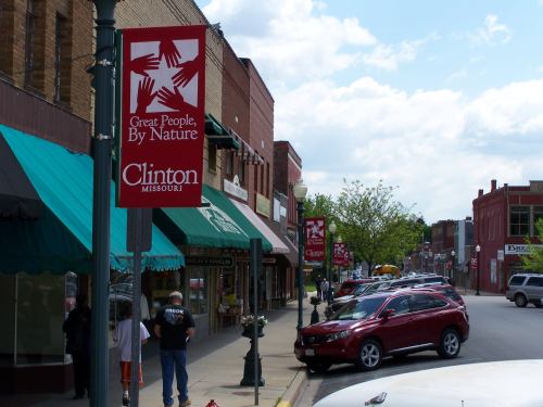 Main street in Clinton, Missouri, featuring banners that read "Great People, By Nature" and shops along the sidewalk.
