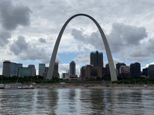 The Gateway Arch stands tall over the St. Louis skyline, reflecting in the river under a cloudy sky.