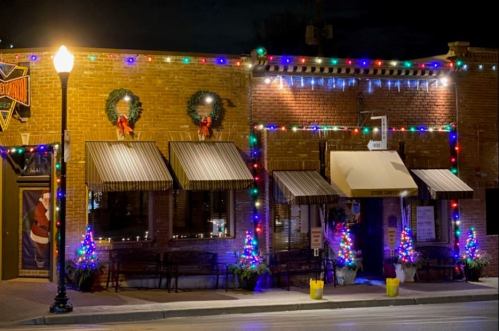 A festive restaurant exterior decorated with Christmas lights, wreaths, and small trees, glowing at night.