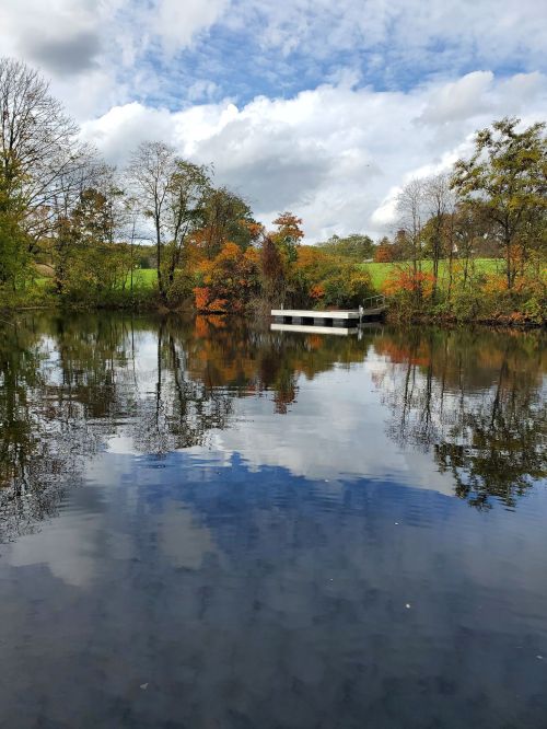 A serene lake reflecting trees and clouds, with a small dock surrounded by autumn foliage.