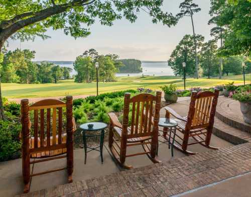 Three rocking chairs on a patio overlook a serene lake and lush greenery under a clear sky.