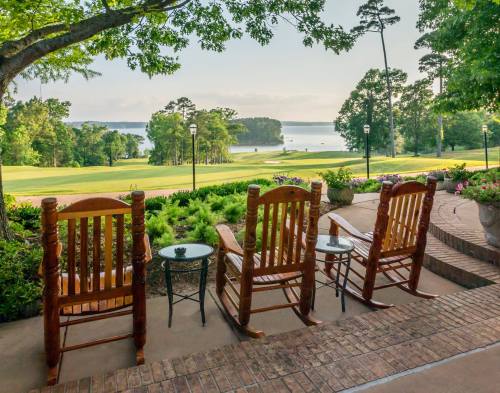 Three rocking chairs on a patio overlook a serene lake and lush greenery under a clear sky.