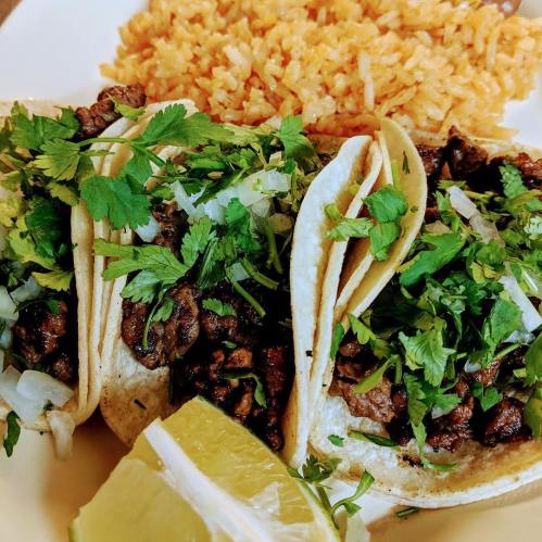 Three tacos filled with seasoned meat, topped with onions and cilantro, served with a side of rice and a lime wedge.
