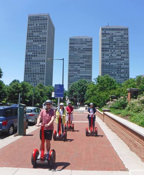 A group of four people riding Segways on a sunny day, with tall buildings and greenery in the background.