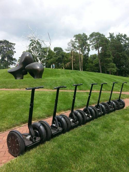 A row of black Segways on a grassy path, with a large abstract sculpture and trees in the background.