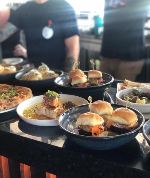 A variety of dishes including sliders, pizza, and appetizers displayed on a table in a bright setting.