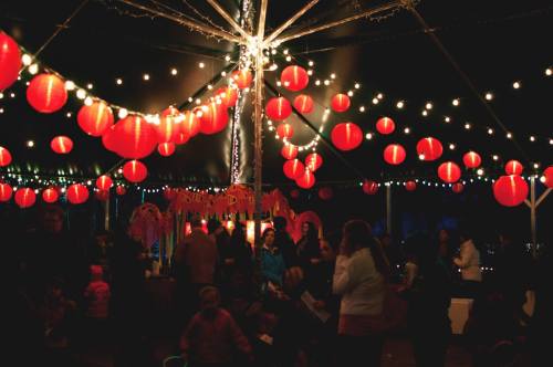 A festive scene with red lanterns and string lights illuminating a crowd under a tent at night.