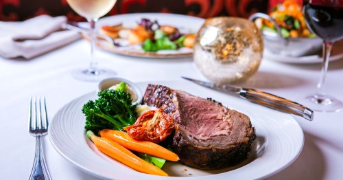 A plated meal featuring roast beef, broccoli, carrots, and a glass of white wine, with a candlelit ambiance.