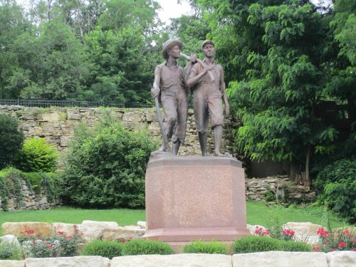 A bronze statue of two boys walking together, surrounded by greenery and flowers in a park setting.
