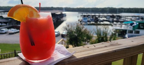 A refreshing red cocktail with an orange slice, overlooking a marina with boats and a scenic waterfront.