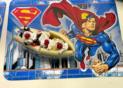 A banana split topped with whipped cream and cherries on a Superman-themed placemat.